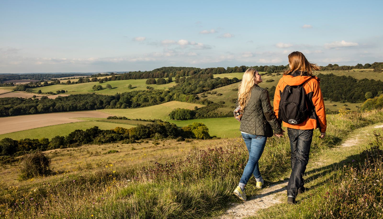 A couple walks along a scenic trail on the South Downs, Hampshire, surrounded by wild grass and overlooking patchwork fields under a bright blue sky.